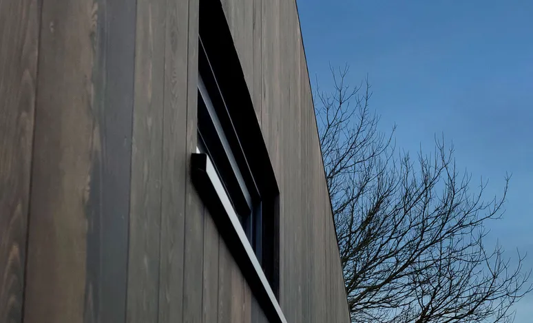 Photograph depicts a building with a minimalist architectural design, featuring black vertical cladding and rosewood panels on its side profile. Small window details add intricate touches. The building, painted white, is accented by a slanted roof. Captured under daylight, the image includes a clear blue sky. In the background, a leafless tree complements the scene, highlighting the stark beauty of architectural photography.