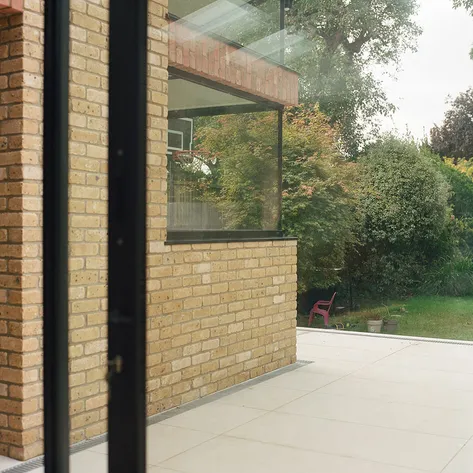 A modern house with brick exterior walls and large glass windows, overlooking a spacious patio and a lush green garden.