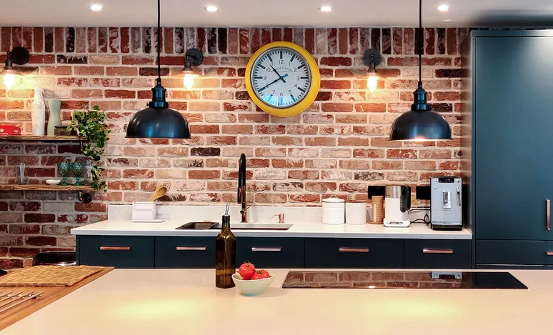 A photo of an industrial style kitchen with a brick wall, black pendant lights and a large yellow clock on the back wall. A white island in front of it with dark blue cabinets and a coffee machine, a paper cup holder, wooden plants on shelves. A white counter top with a red fruit bowl, a glass jar filled with salt and pepper next to the sink, white floor tiles