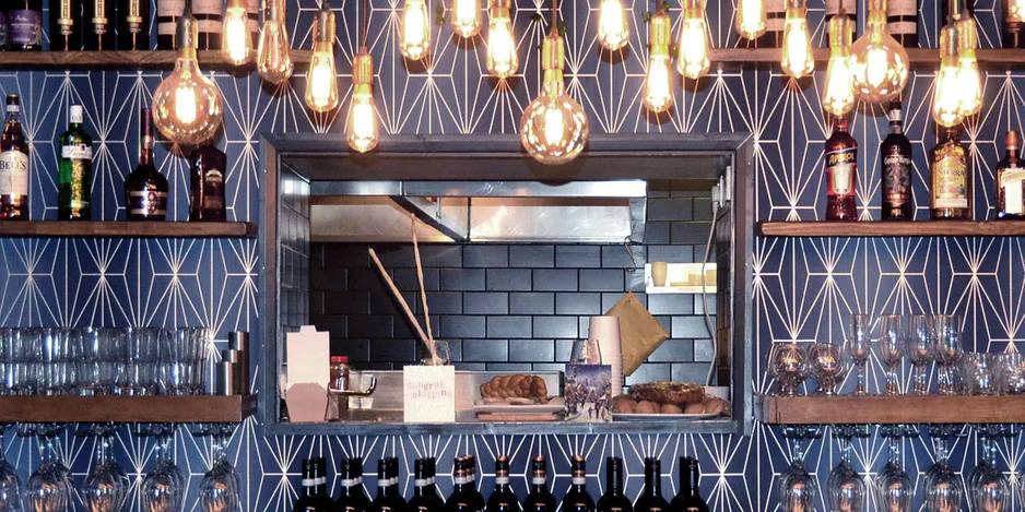 Bar area of the bistro showing the shelves with many bootles and glasses to the sides and a window into the kitchen at the centre. Deep blue tones and colours with brass geometrical patterns.