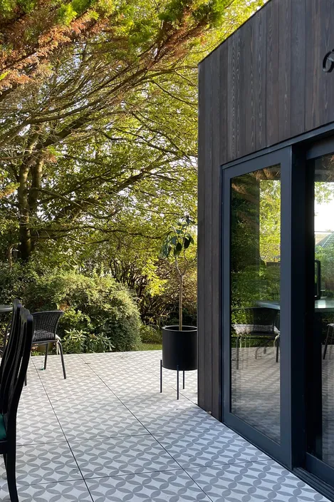 Secluded house extension with dark larch cladding, patio sliding door, and a raised patio with delicate patterned tiles.