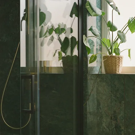 A stylish bathroom with green marble walls, a glass shower enclosure, and a potted plant in a woven basket on the windowsill.