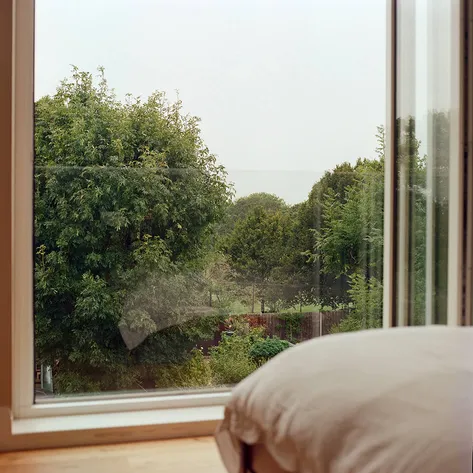 A view of a green landscape through a large bedroom window, with a cozy pillow partially visible in the foreground.