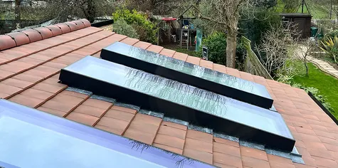 The top view of two flat roof windows on the red tile roof. The focus is set to highlight the contrast between the smooth surface of ceramic tiles against the dark metal beams of each skylight frame