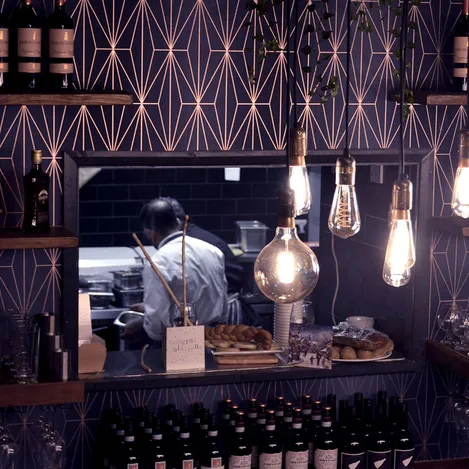 The window into the kitchen from a bar area. You can see the cooks engaged in cooking. Around the window we can see shelves with bottles and a couple of pendant lights framing the view.