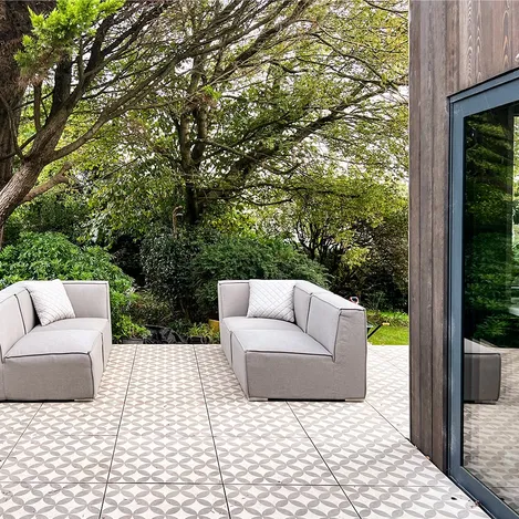 Raised patio with patterned tiles, two grey outdoor sofas, facing green trees in the background.