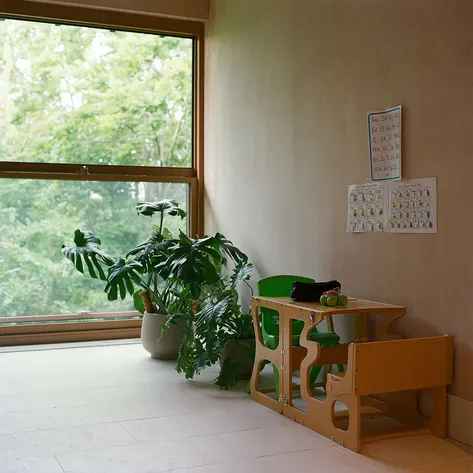 A children's study area with a wooden desk and green chair, next to a large window framing a view of dense greenery outside. 