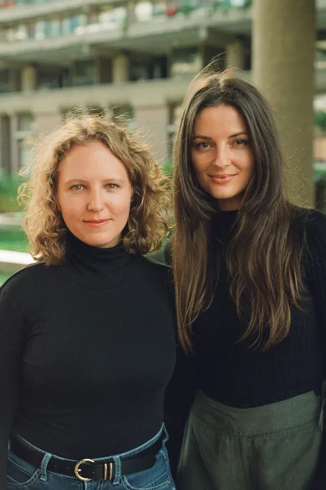 A portrait of two women, both wearing black tops, standing in front of a blurred architectural background.