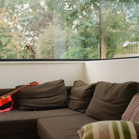 A corner of a modern living room with a brown sectional sofa, a bright orange guitar resting on the cushions, and a large glass window offering a view of a leafy backyard.