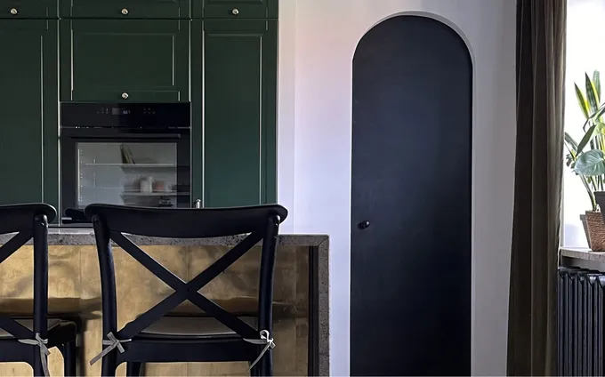 Captured here is a portion of a kitchen that bears the mark of Skala Studio's expertise. With green cabinets, a travertine stone and brass sheet-backed kitchen island, and an arched utility door, the space exudes sophistication. Ample natural light pours in through the window, creating a welcoming atmosphere that combines style and functionality seamlessly.