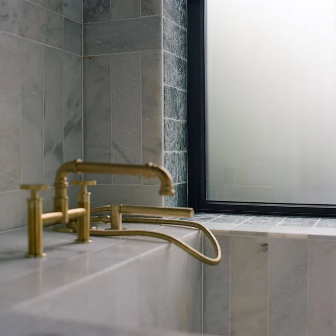 A modern bathroom featuring luxurious marble tiles, with a brass faucet and shower attachment, set against a frosted window that diffuses soft natural light.