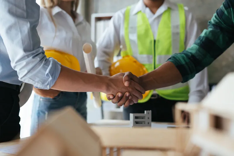 Group of people at conference table, two shaking hands, two holding construction safety helmets.