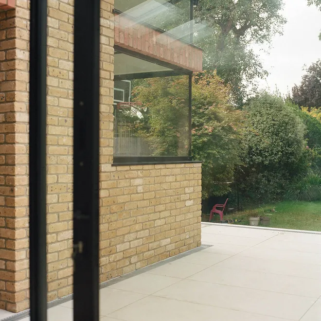 An outdoor patio space with large light stone tiles, adjacent to a brick wall featuring corner windows with expansive glass panes, overlooking a garden filled with greenery.