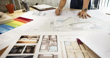 Architect's hand with a pencil annotating on architectural drawing. There is a lot of drawings and colour palettes on the table. They all show simple design layouts and options.