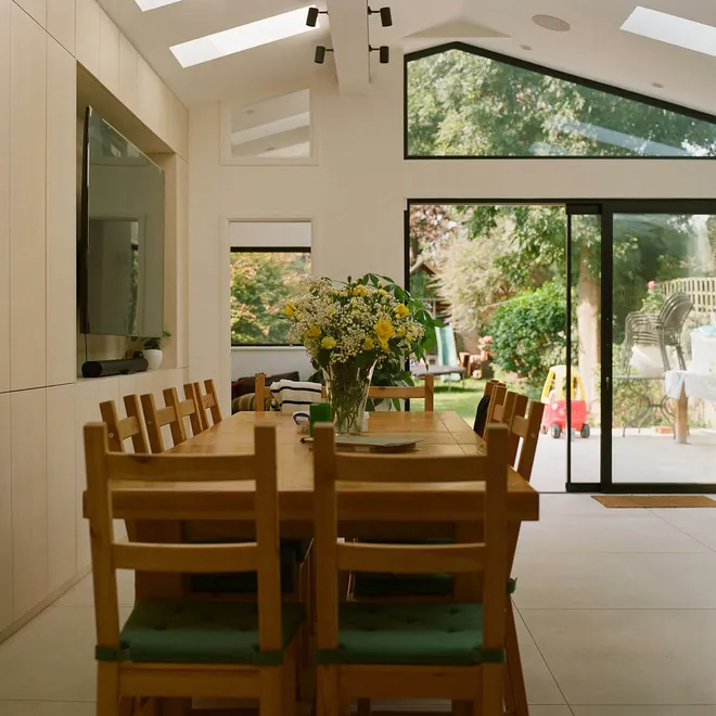 A bright dining room with a long wooden table and chairs, featuring a floral centrepiece, large skylights, and glass doors that open to a lush garden, creating a seamless indoor-outdoor connection.