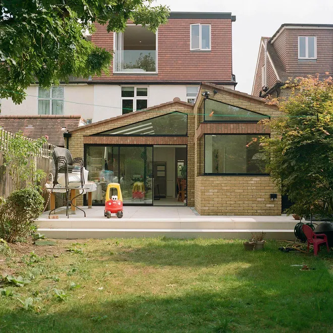 A modern brick and glass extension with large corner windows and sliding doors opens onto a patio and garden, with a children's toy car in front and an open balcony visible on the top floor of the house.