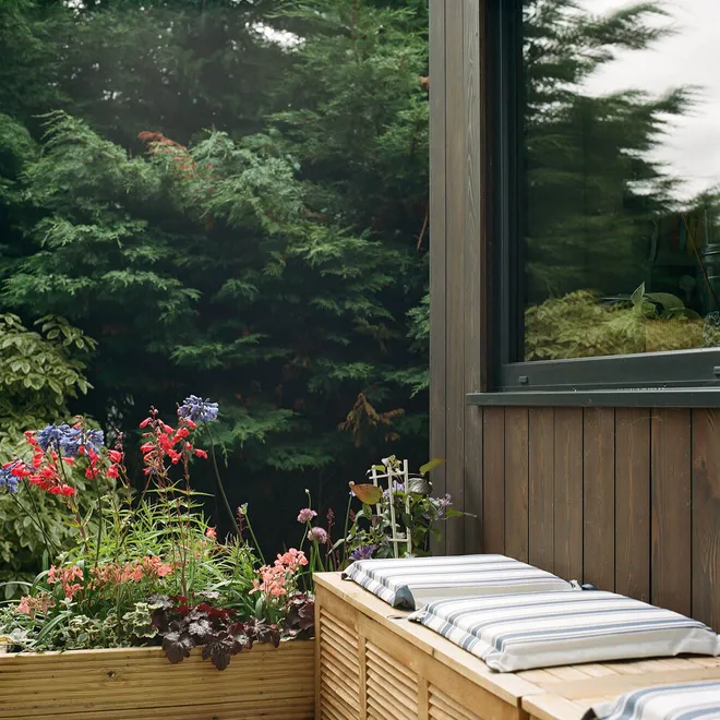 A cosy outdoor garden space with a wooden planter full of colorful flowers, a cushioned bench, and a backdrop of lush green hedges beside a dark wood-clad building.